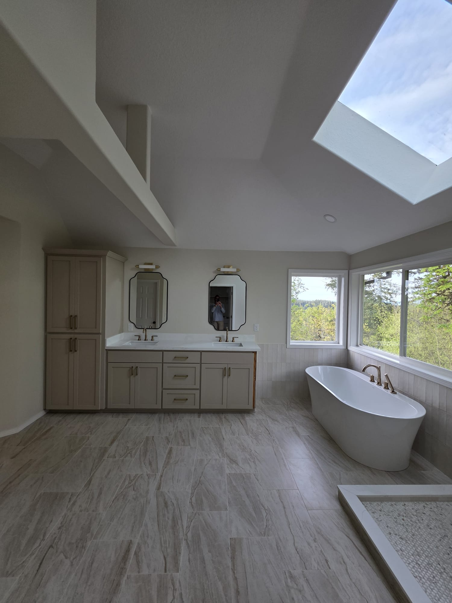 Freshly finished bathroom with vaulted ceiling, skylight, taupe double vanity, and freestanding soaking tub by a large window